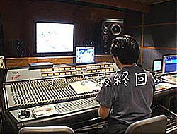 A person sits at a very large desk with various screens, large speakers and a control unit with a vast array of sliders, switches and buttons.